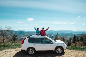 Young couple sitting on the top of the SUV car at mountain peak enjoying the landscape view at summer sunny day. Concept image of “Are SUVs Fit for Off-Roading?” | Outlander Motorsports in Martinez, CA.