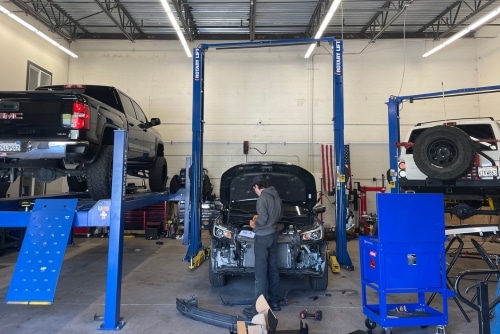 Auto Repair Services in Martinez, CA at Outlander Motorsports. Image of a technician performing preventative maintenance on an open hood car, with a GMC lifted on the left and a white Jeep on the right.