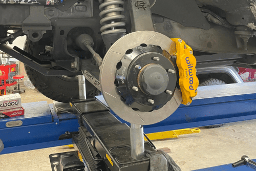 Brake repair in progress at Outlander Motorsports in Martinez, CA. The image shows a lifted vehicle with a close-up of a brake rotor and a yellow Wilwood caliper being serviced on a hydraulic lift.