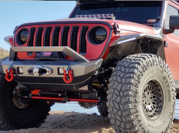 Red off-road vehicle with large tires on sandy terrain near water.